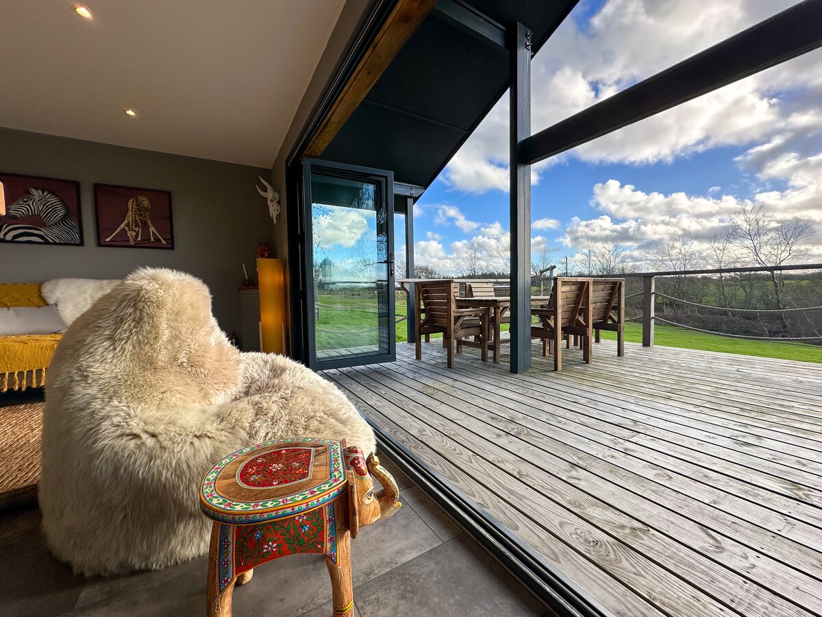 A cozy seating area features a plush, oversized chair with a soft white texture, complemented by a small intricately designed side table. Large bifold doors open to a spacious wooden deck, showcasing serene green views and a dining set for outdoor gatherings.