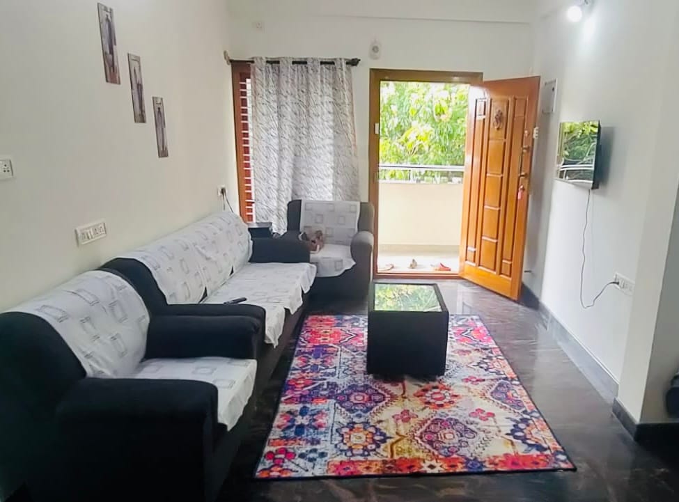 The bright living room features a comfortable seating arrangement, including a stylish black sofa and armchairs. A patterned area rug adds warmth to the space. Natural light floods in from a large balcony door, which opens to a small outdoor area. A wall-mounted television is also visible.