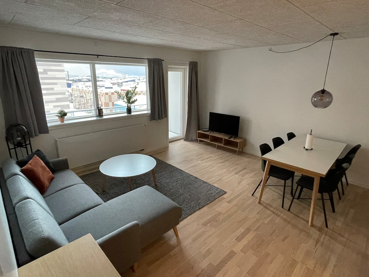 A bright living space is presented, featuring a gray sofa with an orange accent pillow, a round coffee table, and a black television. A dining table with six chairs occupies one side, while large windows allow natural light to illuminate the room.