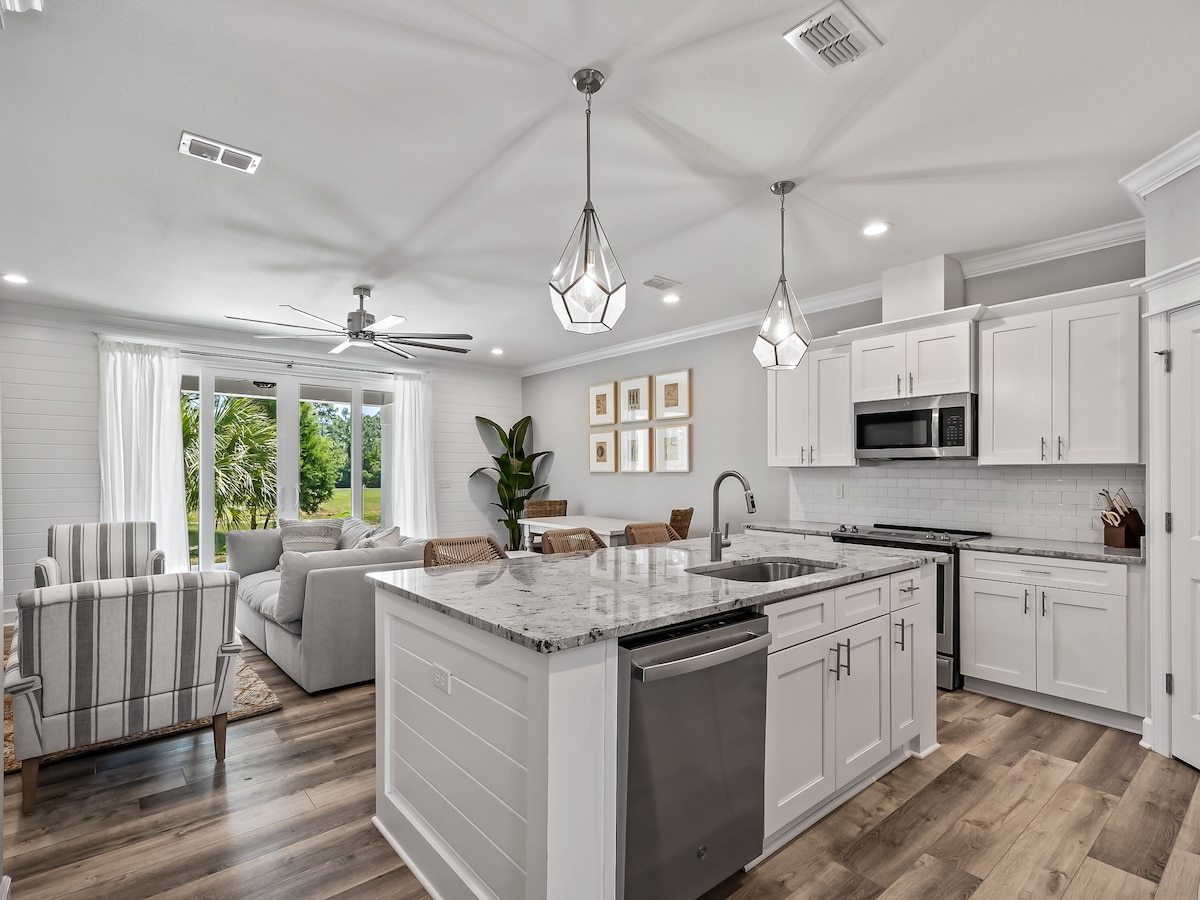 A modern kitchen and living area are displayed, featuring a large island with a marble countertop and stainless steel appliances. Natural light filters through the windows, illuminating the dining space with a wooden table and comfortable chairs. An inviting sofa is visible in the background.