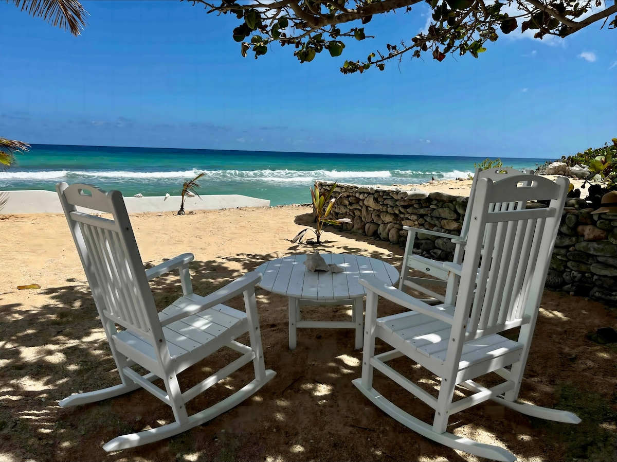 Two white rocking chairs are positioned around a circular table, with a sandy beach and the turquoise waves of the Caribbean Sea visible in the background. The area is shaded by overhead tree branches, creating a serene outdoor space.