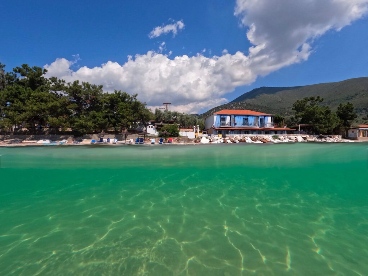 The beachfront area features clear, turquoise waters with a sandy shore. Sun loungers are positioned near the water's edge, while a two-story building is visible in the background, surrounded by trees and hills under a partly cloudy sky.