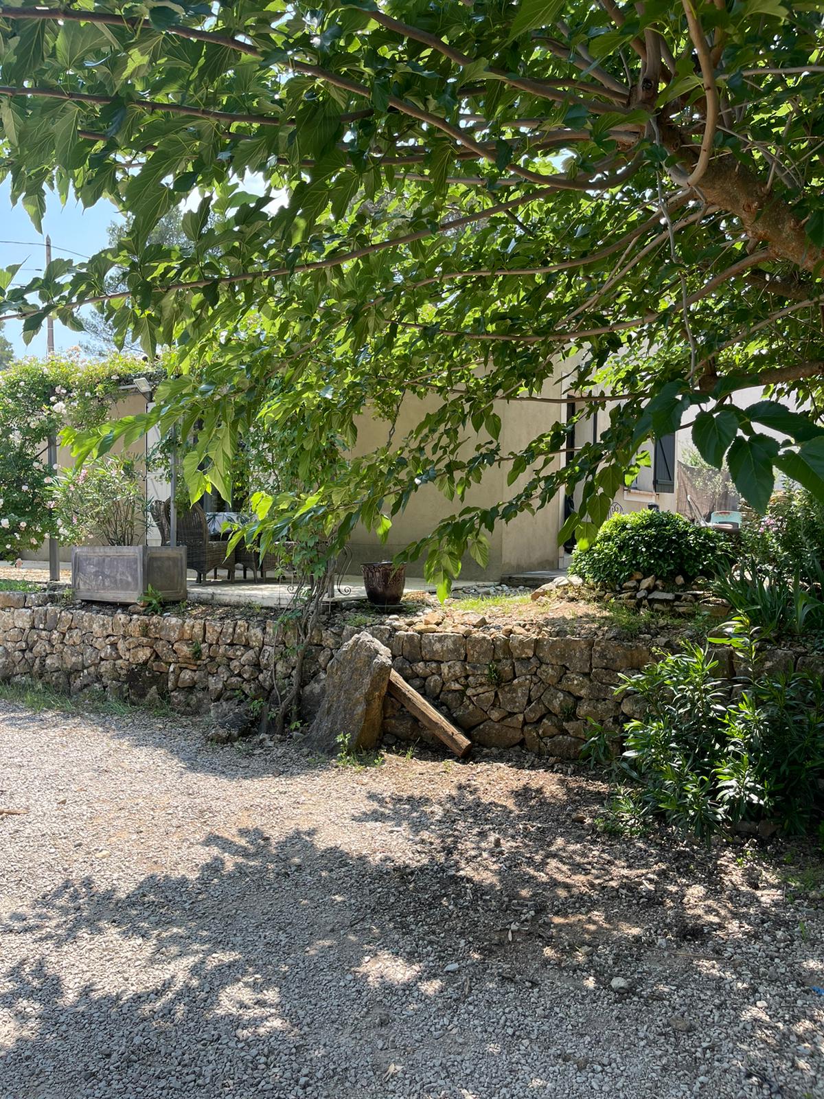 A shaded entrance area is framed by lush greenery and features a stone wall. The gravel path leads toward the house, which is partially visible amid the foliage. Surrounding plants and trees create a serene atmosphere.