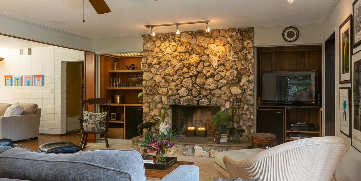 A cozy living area is centered around a large stone fireplace, providing warmth and character. Soft seating options are visible, along with a television and decorative artwork on the walls. Natural light enhances the inviting atmosphere, complemented by tasteful plants and floral arrangements.