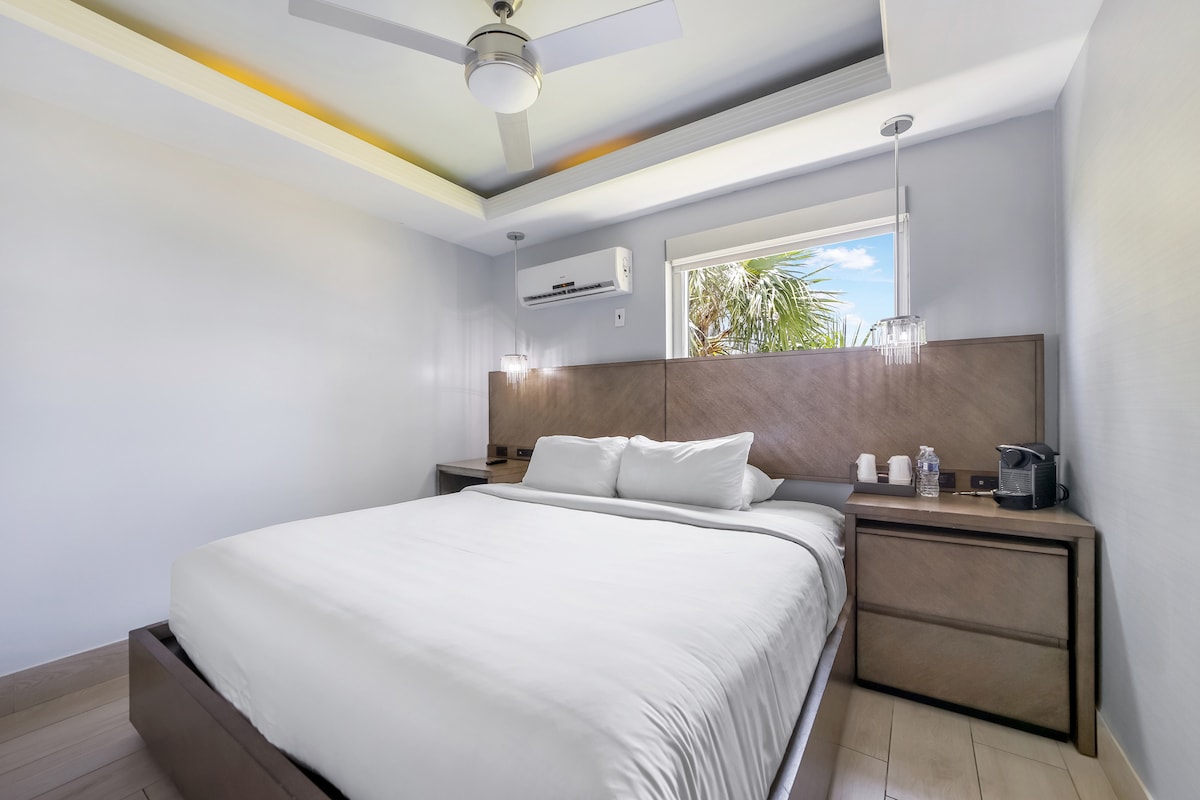 The spacious bedroom features a king bed with crisp white linens, complemented by bedside tables and lamps. Natural light streams through a window, illuminating the light-colored walls and contemporary ceiling fan. A coffee maker and cups are visible on the side table.