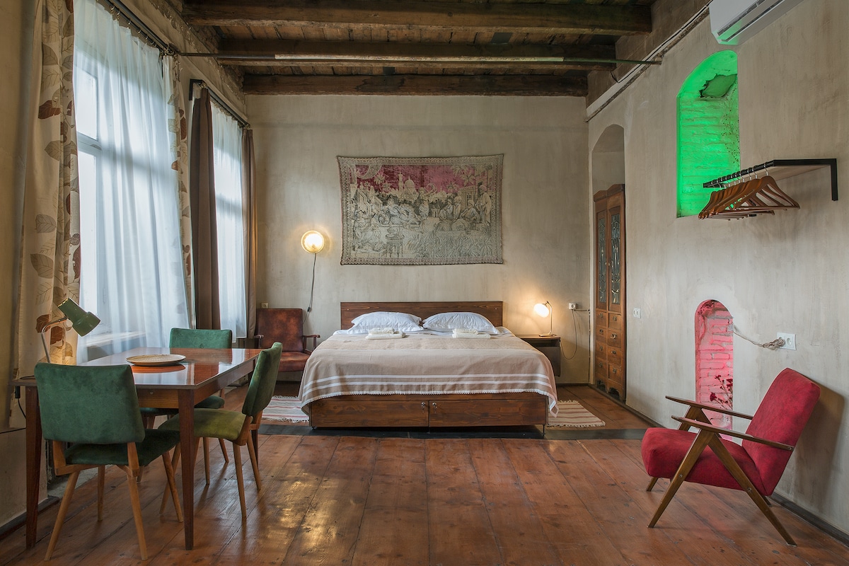 A spacious sleeping area is featured with a wooden bed dressed in hotel-quality linens. Colorful chairs surround a wooden table, and natural light filters through large windows, complemented by understated wall art and exposed wooden beams overhead.