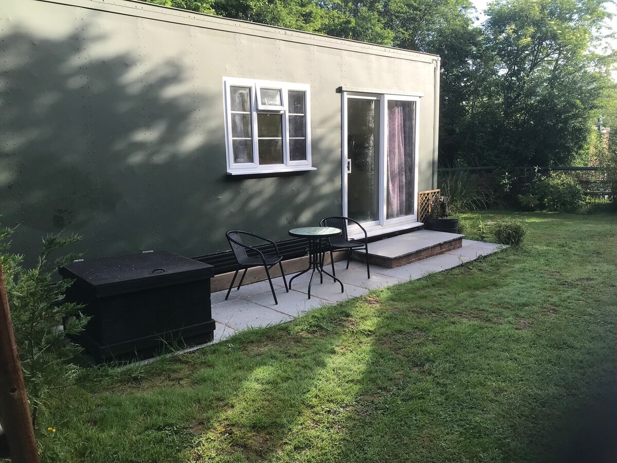 A small outdoor seating area is visible, featuring a round table surrounded by two black chairs. The entrance to the space is highlighted by a sliding glass door, with natural light filtering in. Lush greenery surrounds the area, adding to a peaceful ambiance.