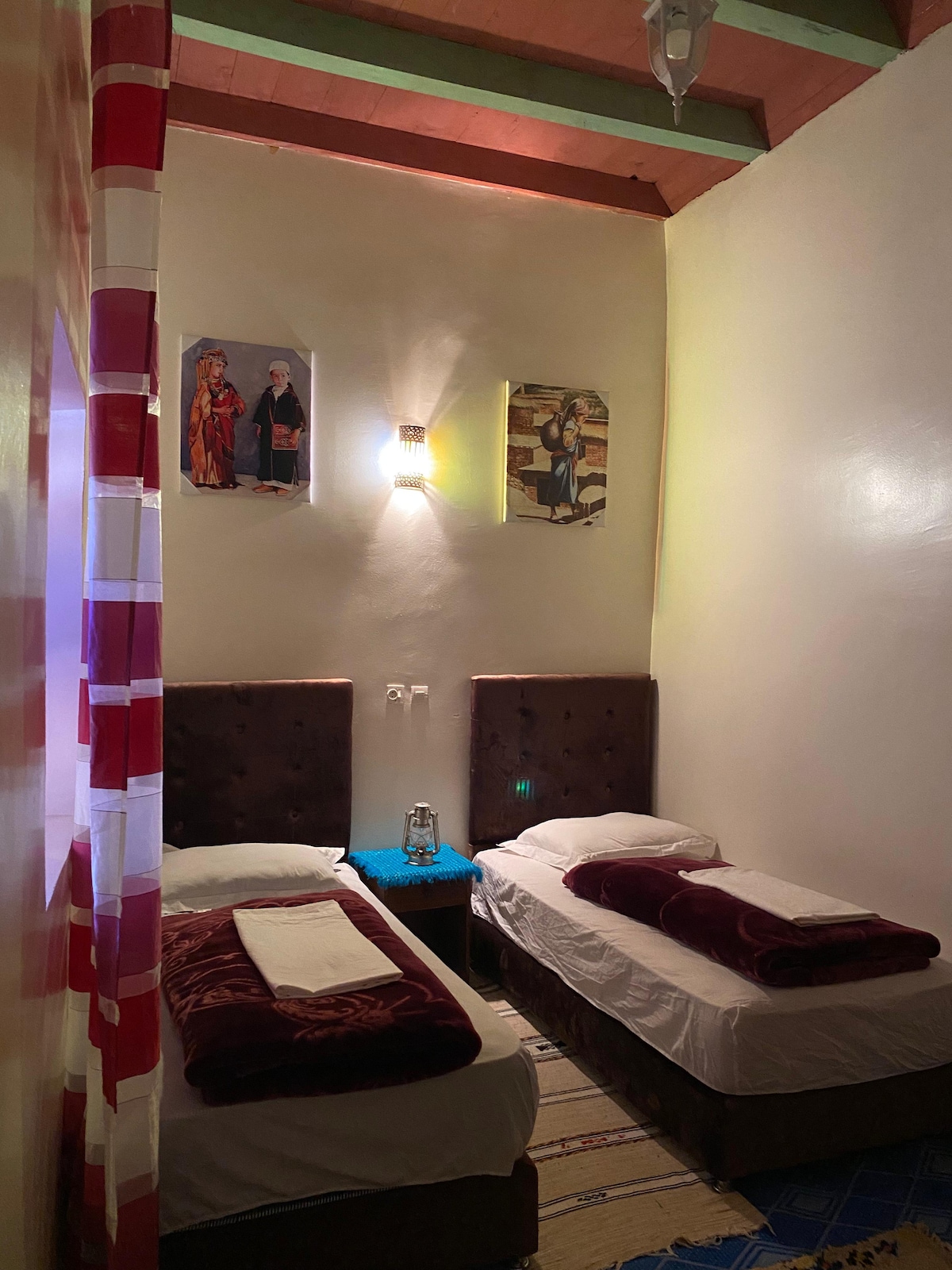 A cozy guest room features two spacious beds with soft linens and maroon blankets. Colorful artwork adorns the walls, complemented by warm lighting from a stylish wall fixture. A small table is positioned between the beds, adding functionality to the space.