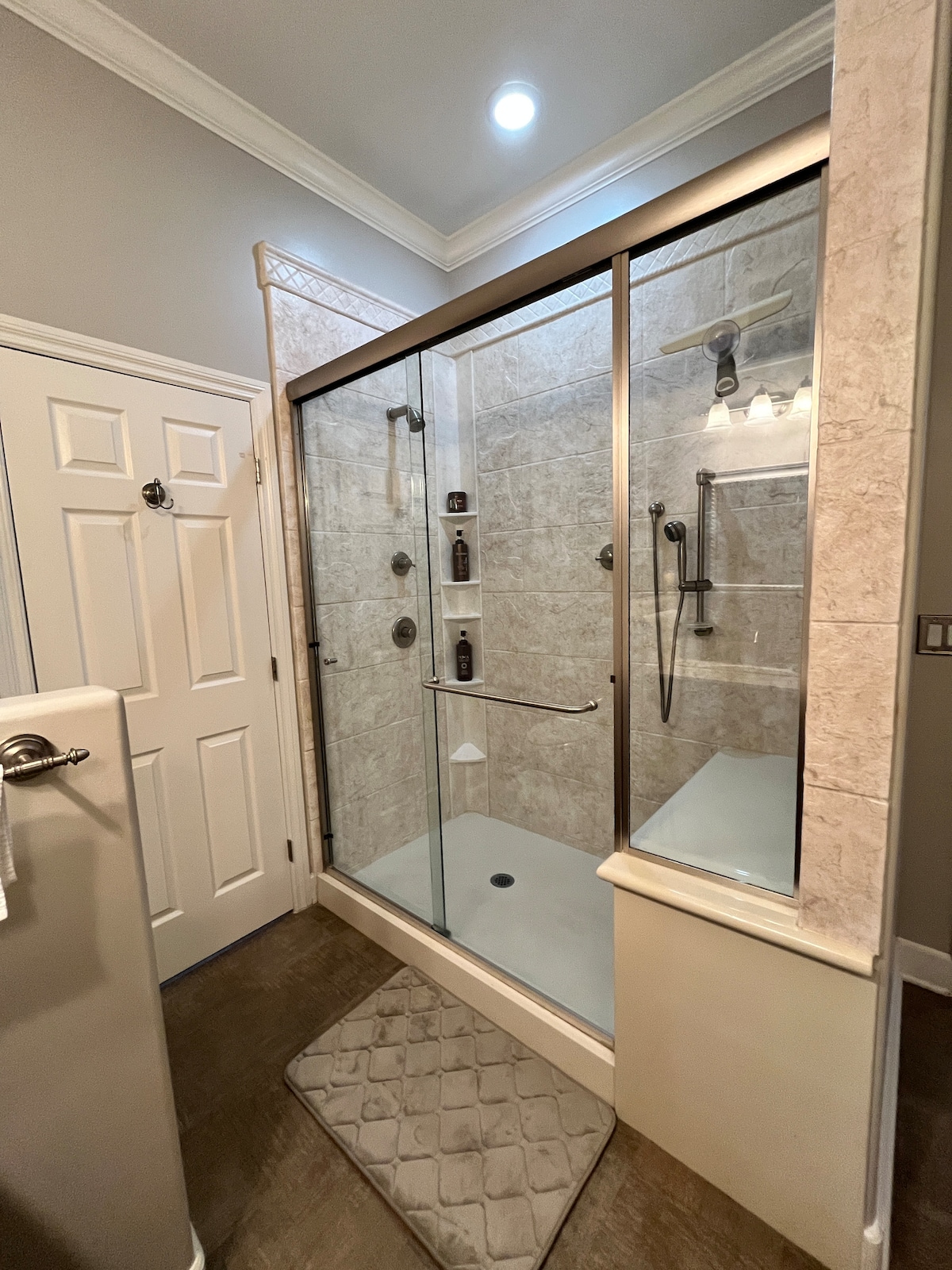 A spacious glass shower is enclosed with modern framing, featuring dual shower heads and built-in shelving for toiletries. A plush mat lies on the floor outside the shower, and soft lighting illuminates the area. Neutral tones grace the walls, enhancing the clean and contemporary look.