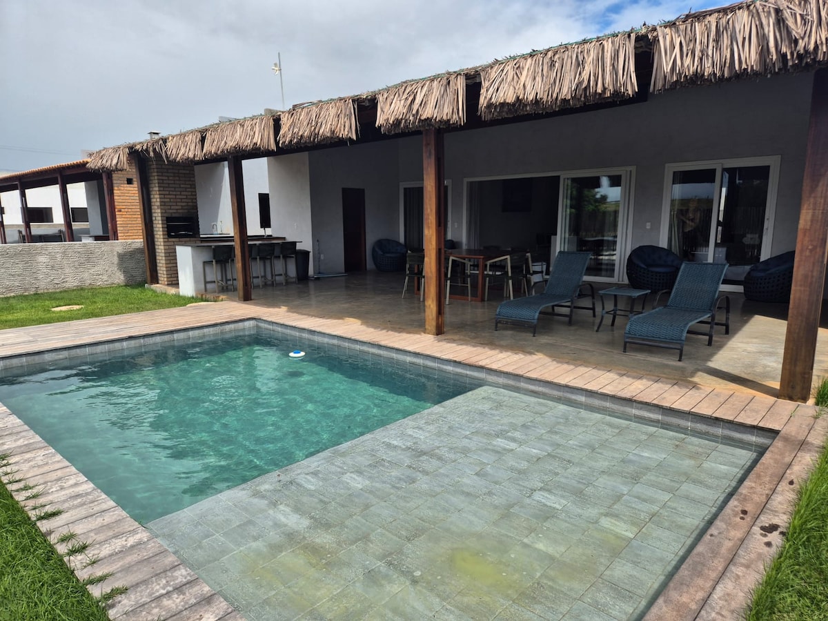 A modern house is depicted with a private pool featuring a shallow area for loungers. A covered terrace with thatched roofing offers shaded seating. The pool area is surrounded by well-maintained grass, and large glass doors reveal a spacious interior connecting outdoor and indoor spaces.