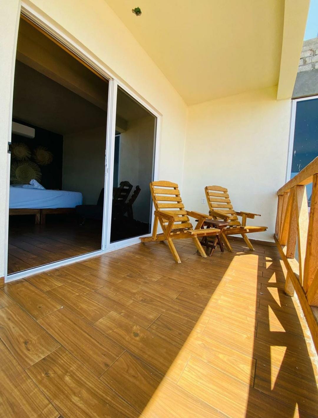 A small balcony features two wooden chairs positioned beside a wooden table. An expansive glass door leads to the interior, allowing natural light to illuminate the space. The floor displays a warm wood finish, contributing to a comfortable outdoor setting.