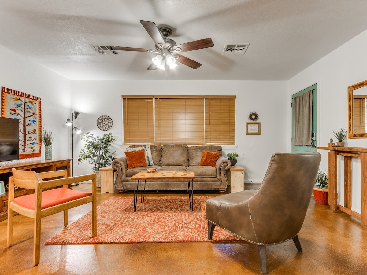 The great room features a comfortable seating arrangement, including a queen sofa bed and two accent chairs. A coffee table sits in the center, surrounded by plants. Natural light enters through blinds, and a ceiling fan adds functionality to the space.
