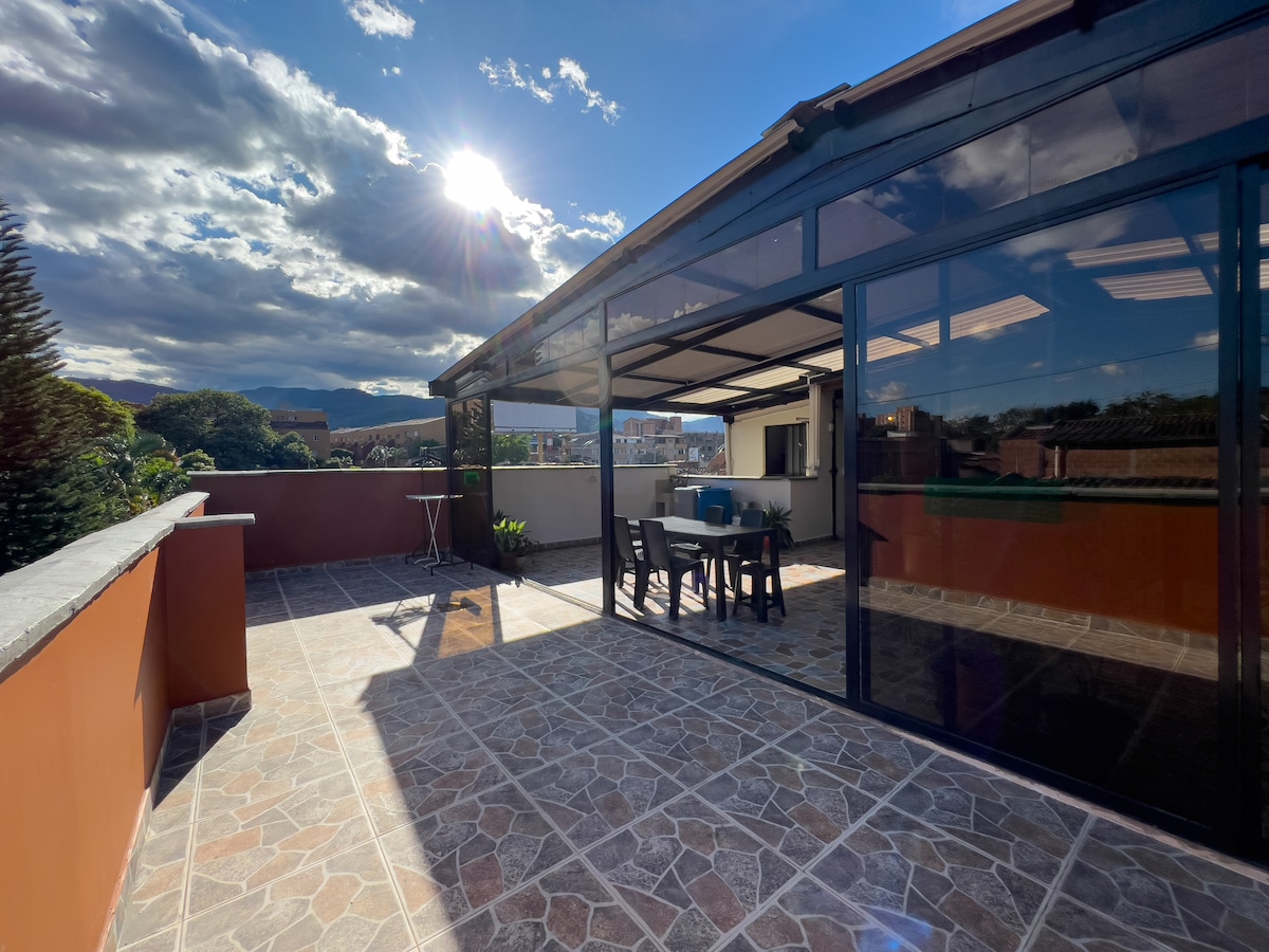 A spacious terrace is seen, featuring a table and chairs under a modern awning. Large glass doors provide views of the surrounding landscape, while sunlight floods the area. Natural light reflects off the tiled floor, enhancing the open atmosphere.