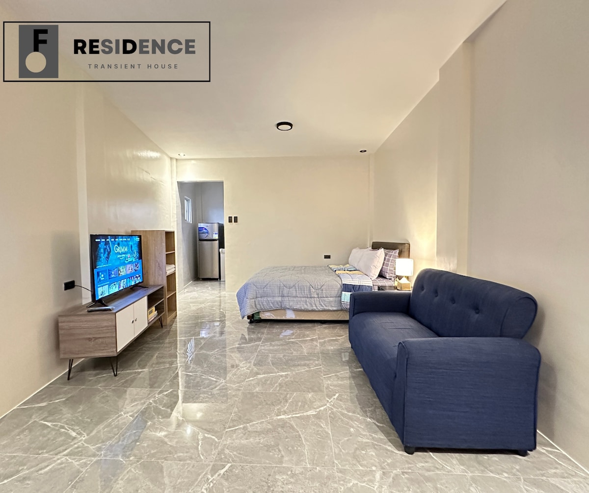 A spacious room features a comfortable blue sofa and a neatly made bed with soft linens. A flat-screen TV sits on a modern console against the wall, with light reflecting off the glossy floor tile. An open doorway leads to a kitchenette.