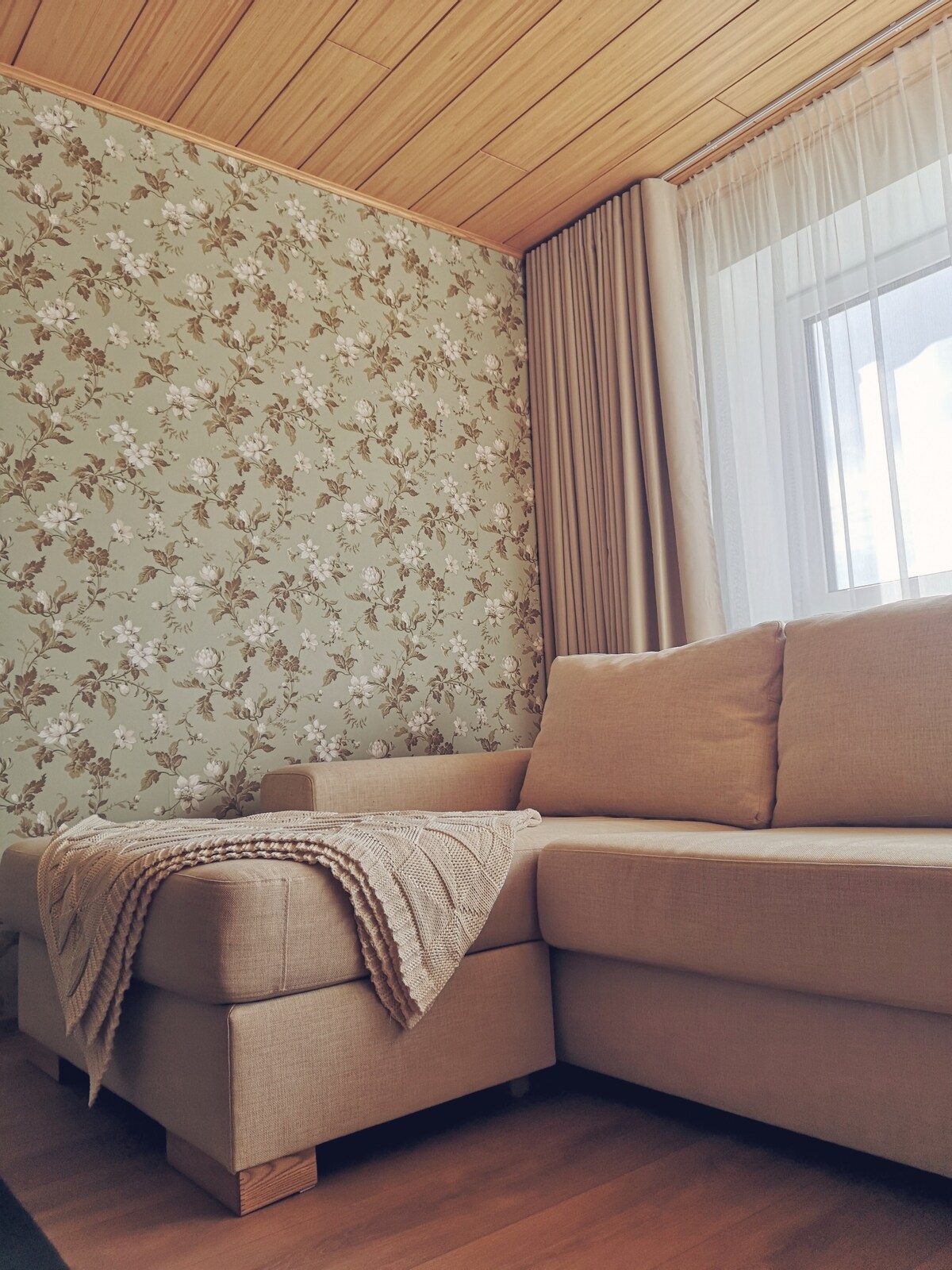 A modern living area features a light-colored sectional sofa with a soft throw draped over it. A floral patterned wall adds a touch of nature, while large windows allow natural light to illuminate the space.