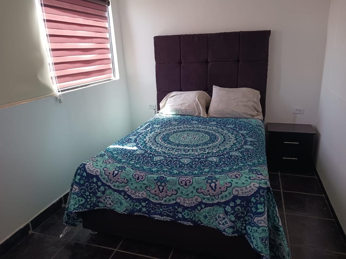 A spacious bed is dressed with a colorful patterned blanket and complemented by two pillows. A bedside table is positioned next to the bed, and natural light enters through a window covered with horizontal blinds.