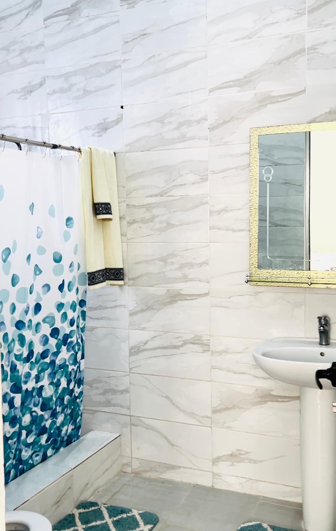 A modern bathroom features light-colored marble wall tiles and a white sink. A decorative shower curtain with a blue and green design adds a pop of color. A large mirror with an ornate gold frame reflects the space, while a plush bath mat is placed on the floor.