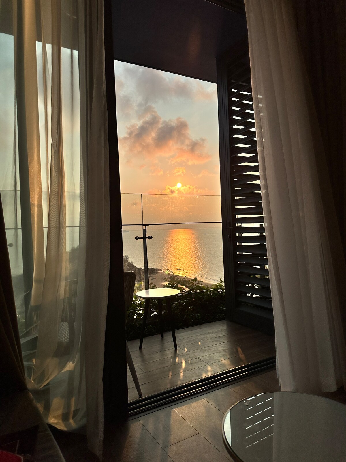 A serene view is visible through sheer curtains, revealing a sunset over the water. The reflection of the vibrant sky on the calm surface is complemented by a small balcony with a table and chair, enhancing the connection to the serene outdoors.