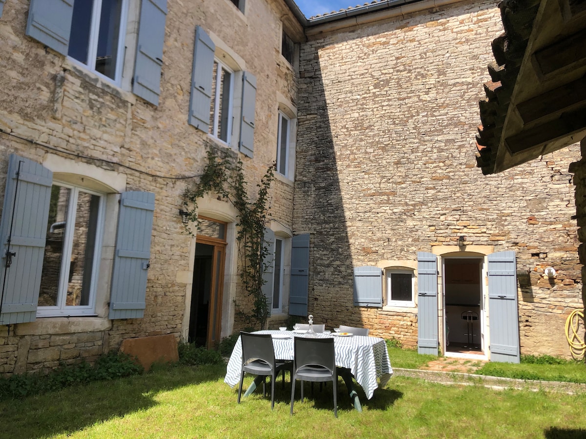 A serene outdoor courtyard is featured, surrounded by stone walls and rustic blue shutters. A table is set for dining, accompanied by four chairs, all placed on a grassy area. Bright sunlight filters in, creating a welcoming environment for relaxation.