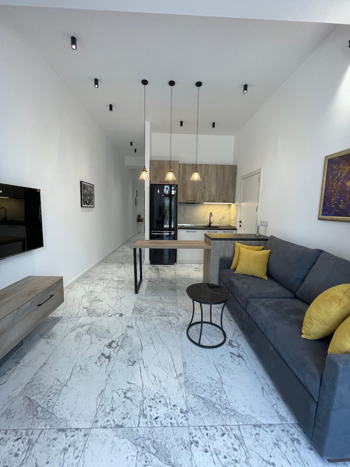A modern studio interior is depicted, featuring an open layout with a sleek kitchen and living area. Pendant lights hang over a wooden dining table, while a gray sofa accented with yellow cushions adds a pop of color. Light marble flooring enhances the airy ambiance.