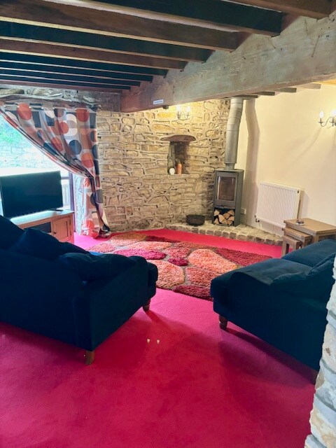 A welcoming living area is characterized by exposed beams and stone walls. A wood-burning stove is situated against a stone feature, complemented by a cozy area rug. Colorful drapes frame the window, while inviting seating arrangements create a comfortable space for relaxation.