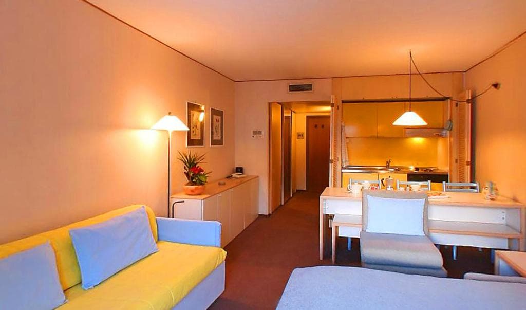 A well-appointed studio apartment features a light-colored sofa complemented by a grey bed. A dining area with a white table and light chairs is visible, alongside a kitchenette equipped with overhead lighting and essential appliances. Soft lighting enhances the welcoming ambiance.