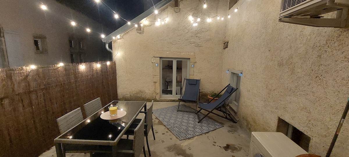 A cozy terrace is illuminated by warm string lights, creating a welcoming ambiance for evening relaxation. A glass table and two lounge chairs are positioned on a concrete surface, surrounded by privacy fencing. Double doors provide access to the indoors, enhancing the outdoor living experience.