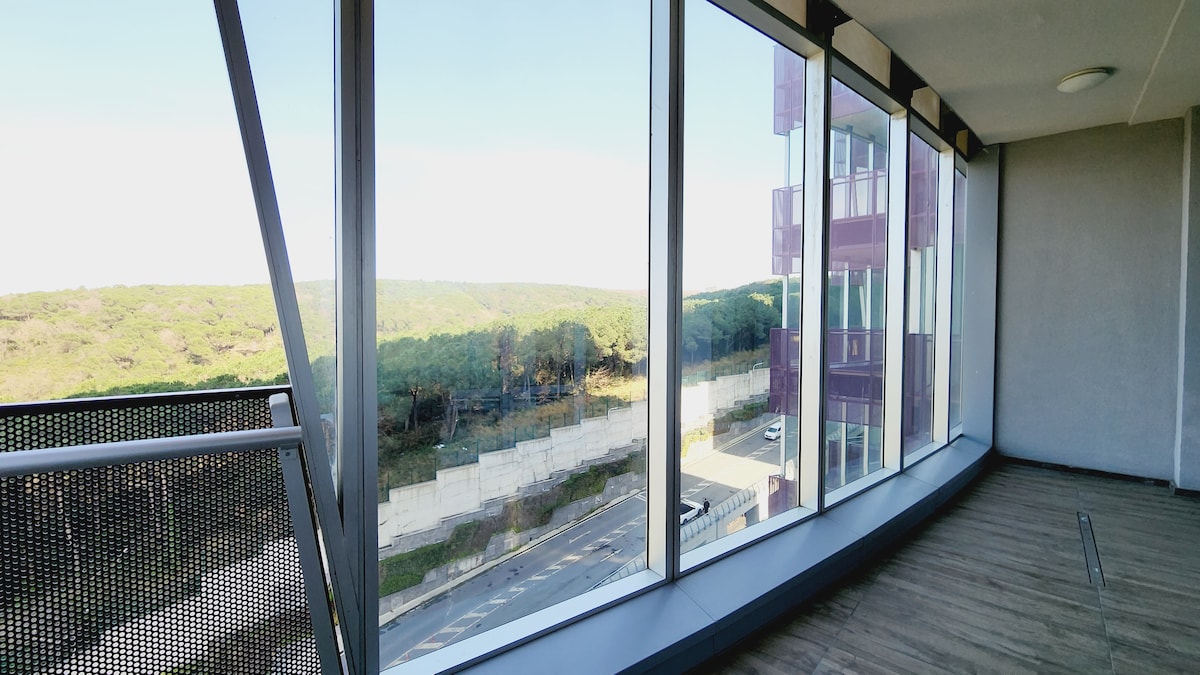 A spacious terrace is showcased, featuring large glass windows that offer an unobstructed view of the surrounding greenery. The wooden flooring enhances the modern feel, and natural light fills the space, creating a bright and open atmosphere.