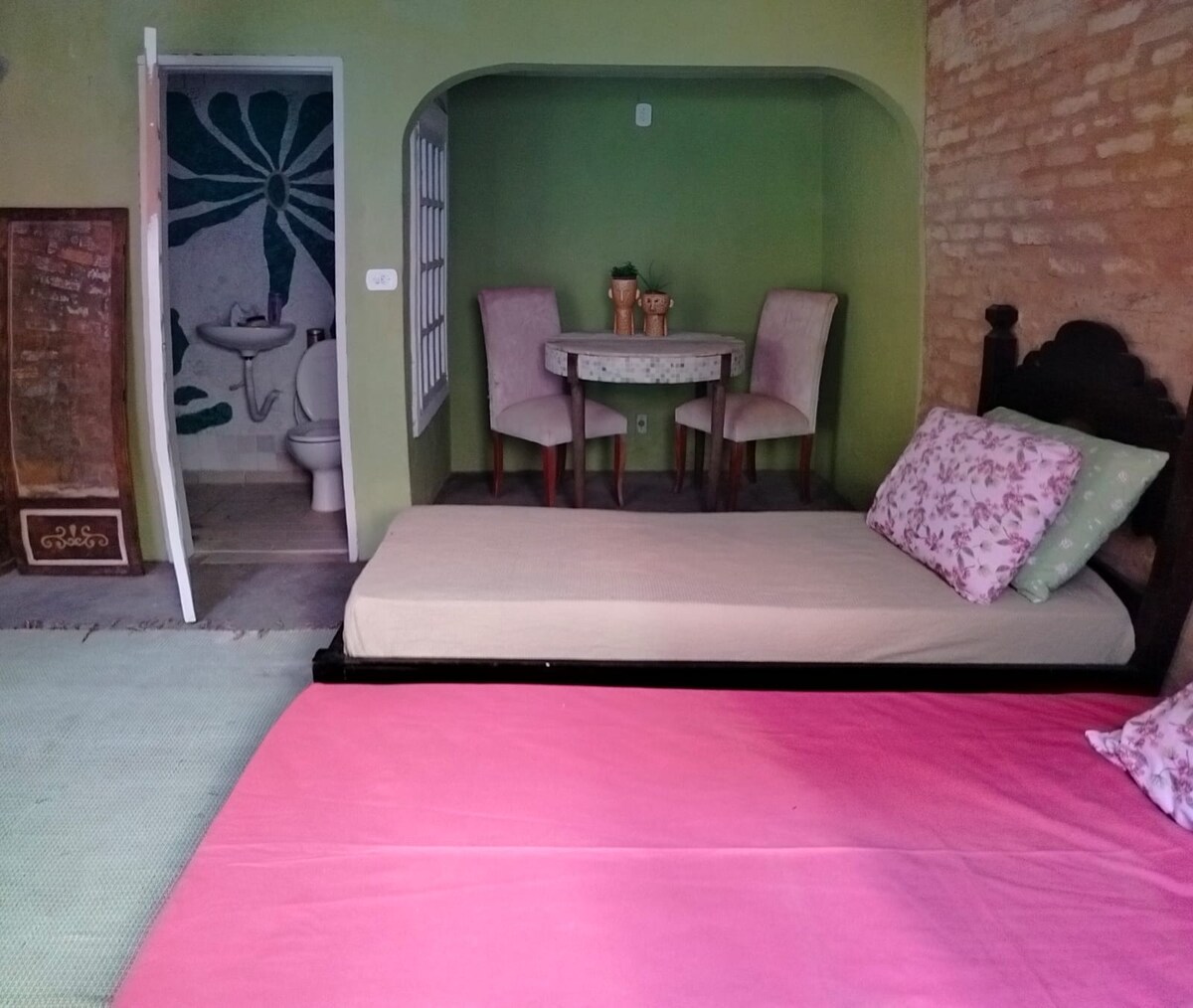 An inviting sleeping area features two beds, one in pink and one in a neutral tone, adorned with floral-patterned pillows. A small dining table with two upholstered chairs is seen in the background, complemented by a green wall and a partially visible bathroom.