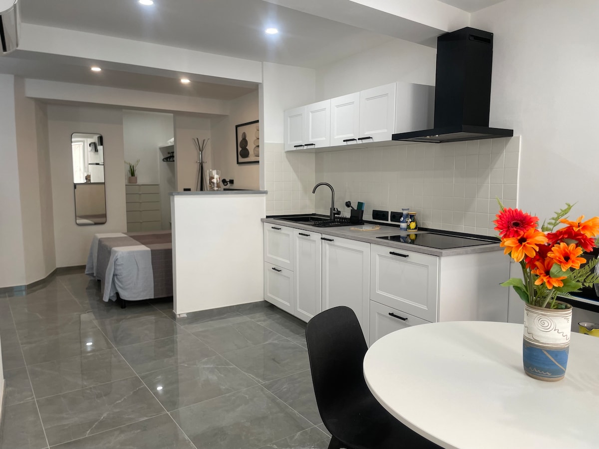An open-concept space reveals a modern kitchen with white cabinetry and dark countertops. A round table with a black chair sits adjacent to the kitchen. A comfortable bed with light bedding is visible in the background, complemented by a vibrant bouquet of flowers on the table.