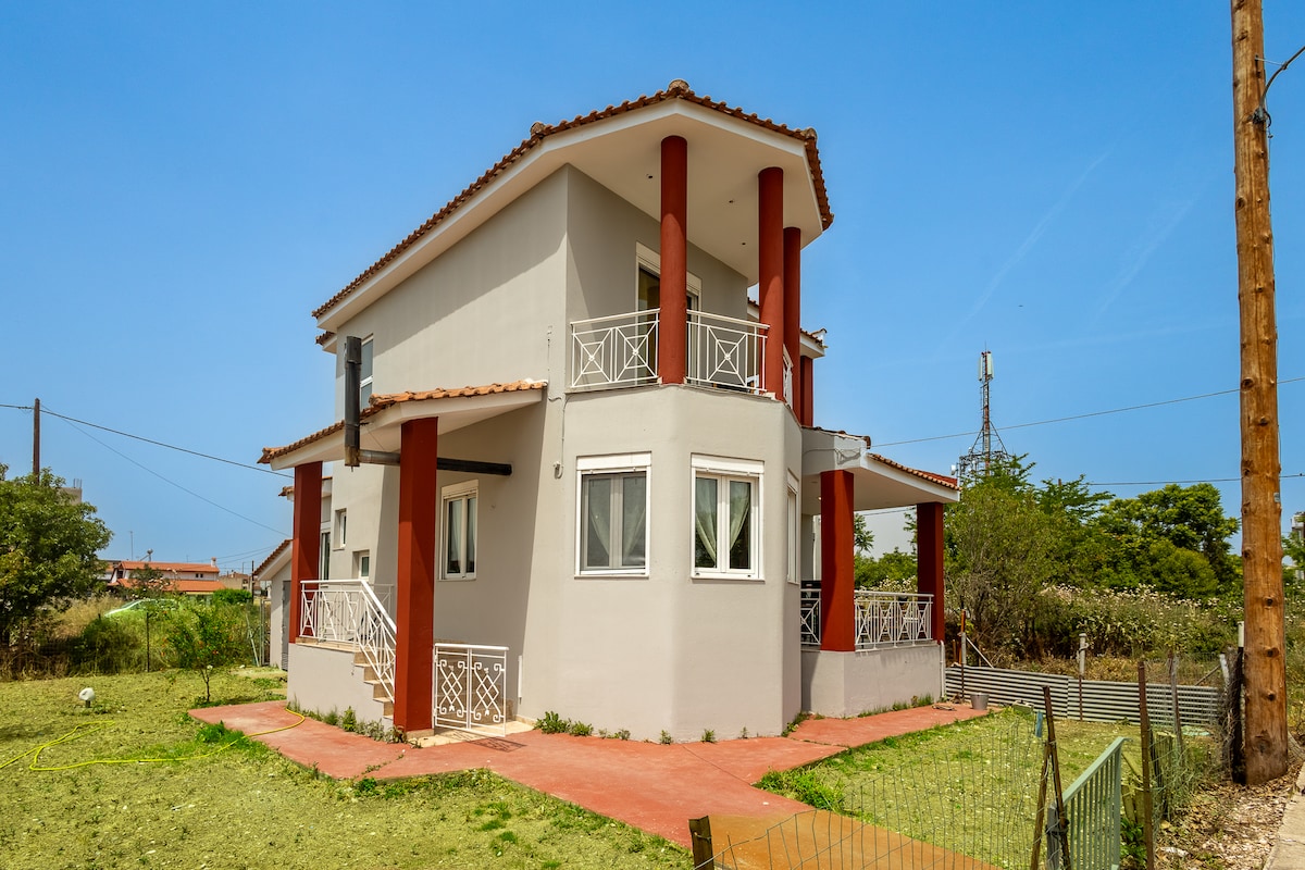 The two-story house features a modern architectural design, with a light exterior and striking columns at the entrance. Windows allow ample natural light, and a spacious yard surrounds the property, providing an open and airy atmosphere.
