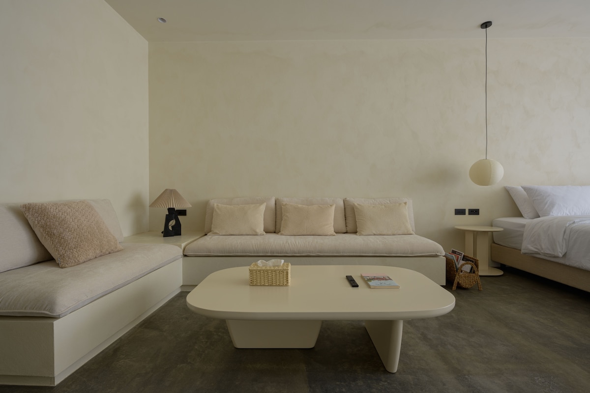 The room features a spacious seating area with a custom-designed sofa adorned with soft cushions. A minimalist coffee table sits at the center, accompanied by a basket. The bed is positioned in the background, while a pendant light provides gentle illumination.