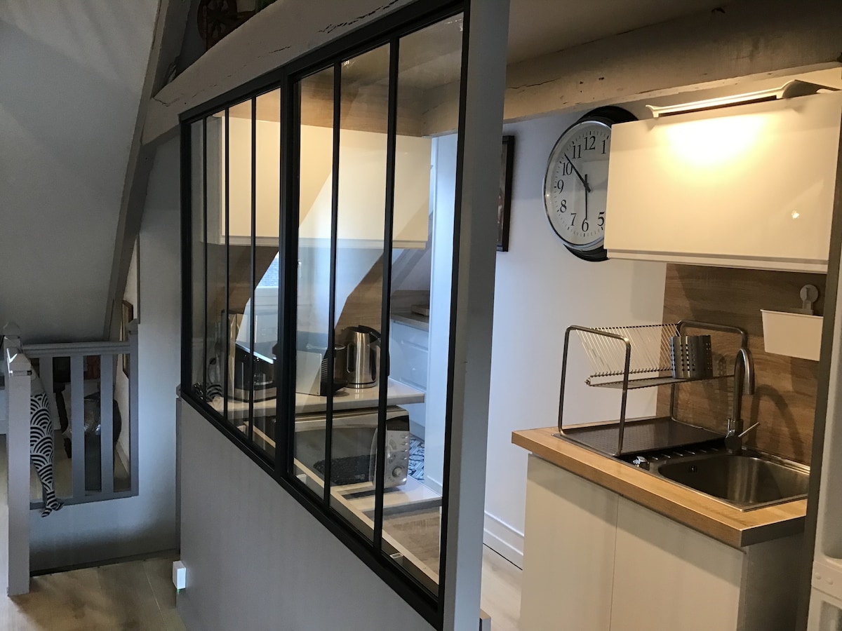 A modern kitchen area is separated by a glass partition, featuring a sleek countertop and a stainless steel sink. Appliances include a kettle and a microwave, while a wall clock adds a functional touch. Natural light filters through the glass, brightening the space.