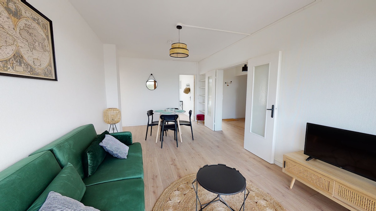 A bright living area showcases a green sofa with decorative pillows, accompanied by a small black coffee table. A round rug lays beneath the table, and a dining table with chairs is visible in the background, beside large windows that allow natural light to fill the space.