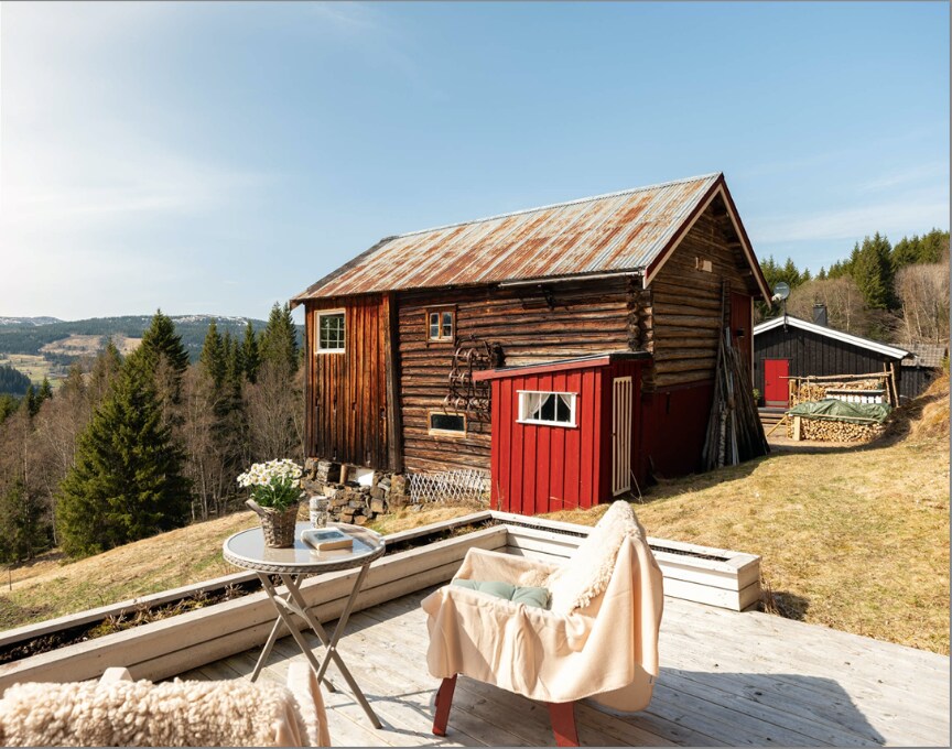 An aged wooden cabin displays a rustic charm, set against a backdrop of green trees and distant hills. A patio area features a small round table and a wooden chair, inviting guests to enjoy the peaceful surroundings.