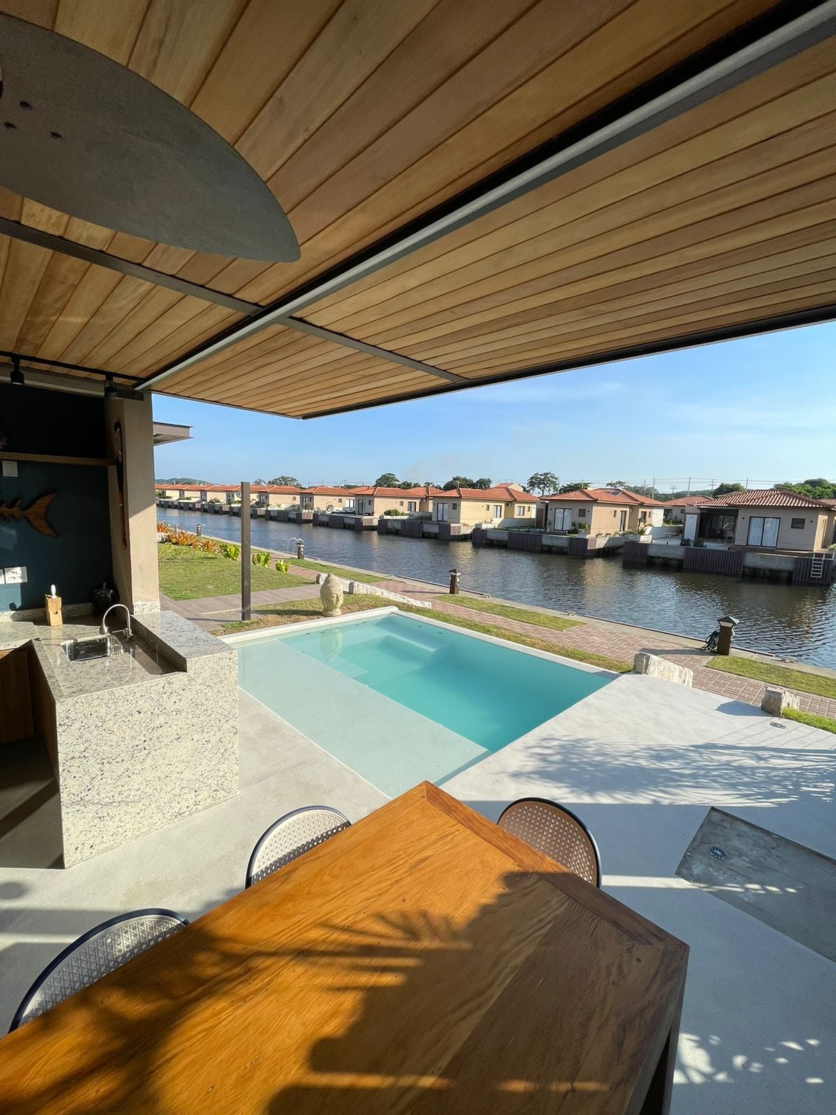 An outdoor dining area features a wooden table and metal chairs adjacent to a clean, rectangular pool. The pool overlooks a tranquil canal lined with charming houses. A covered area provides shade, while a well-equipped outdoor kitchen is visible nearby.
