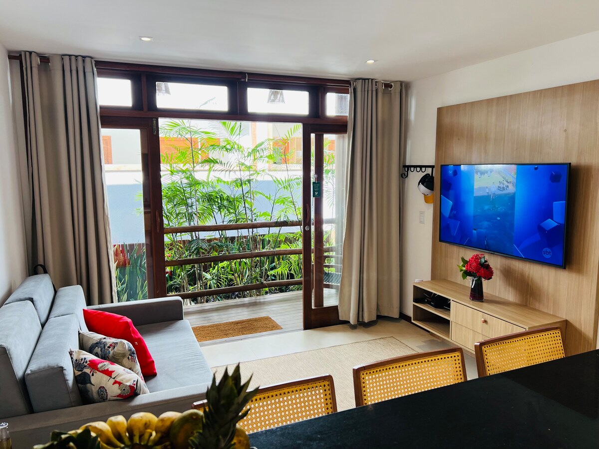 A modern living area features a comfortable gray sofa with colorful cushions, positioned near large windows that provide views of lush greenery. A flat-screen TV is mounted on a wooden panel, and a dining area is visible in the foreground, enhancing the open layout.