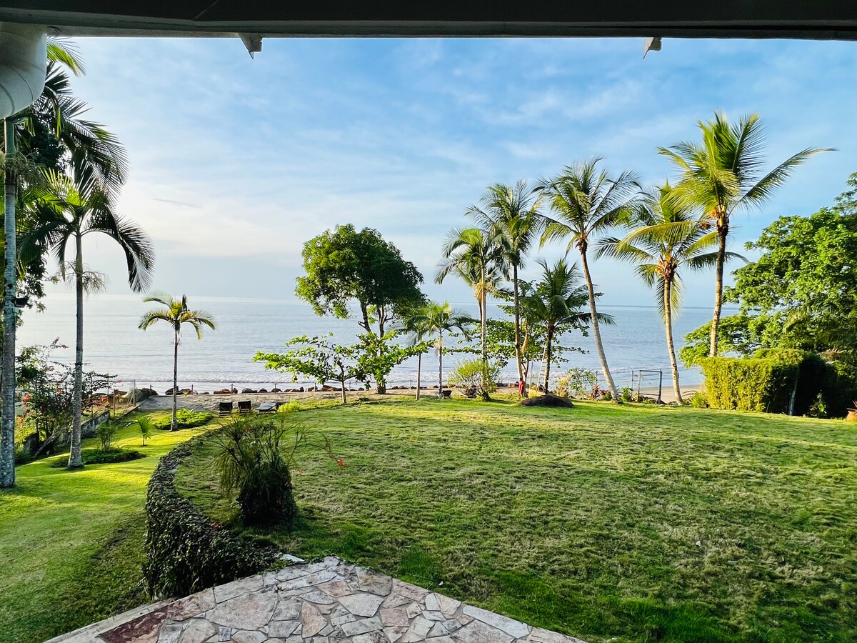 A vast garden area showcases lush greenery and palm trees, leading to a serene beachfront. The calm sea is visible in the distance, while the sky displays soft blue hues. Well-maintained grounds offer a tranquil setting for relaxation.