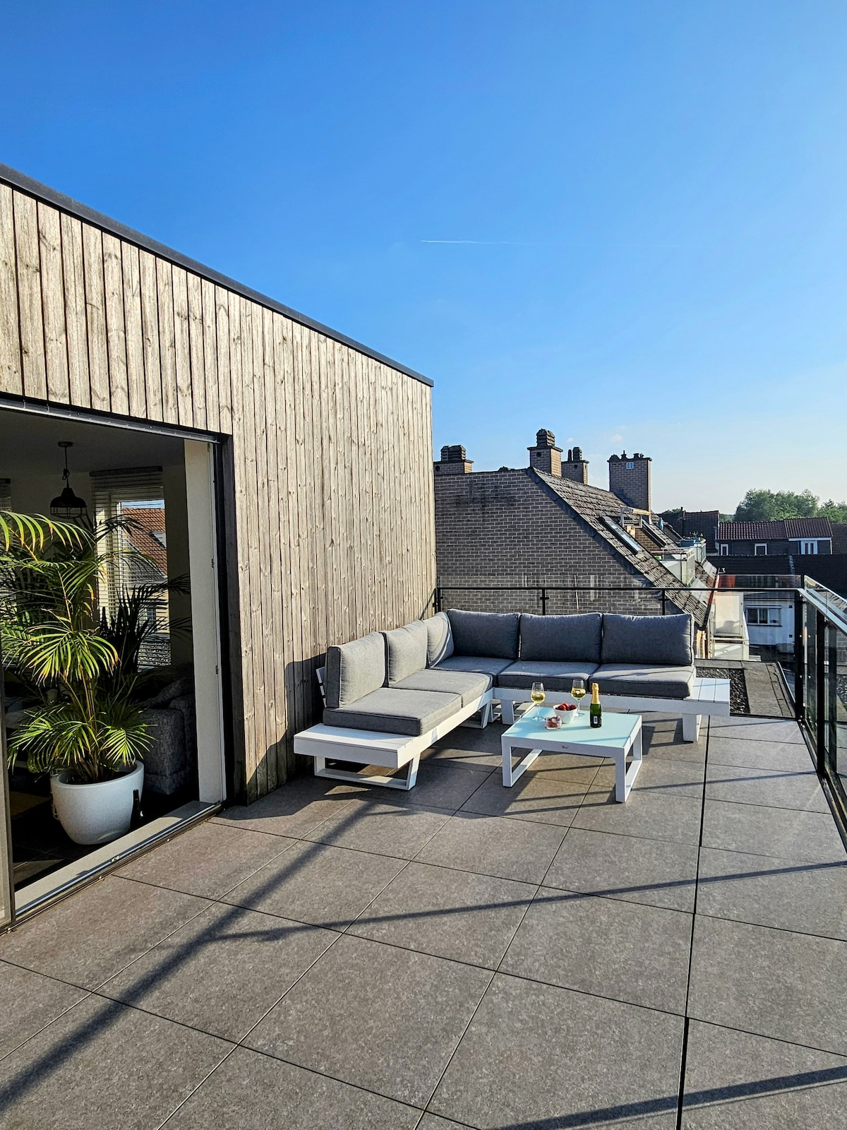 A spacious balcony offers comfortable outdoor seating with a sectional sofa and a coffee table. Sunlight illuminates the area, highlighting the modern design. Surrounding views of rooftops are visible, presenting a serene atmosphere for relaxation.