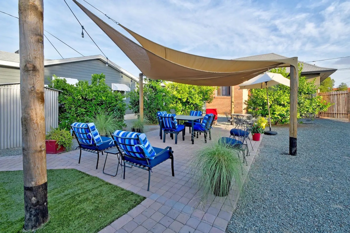 An outdoor seating area features a large shade sail supported by wooden posts. Comfortable lounge chairs with blue and white stripes are arranged around a central table, surrounded by greenery and potted plants. The area is well-defined by paved and gravel pathways.