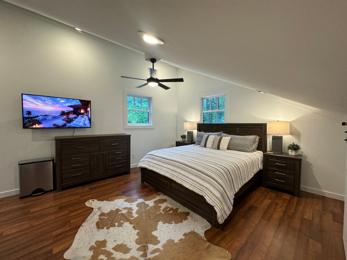 A spacious bedroom is presented with a king size adjustable bed, dressed in neutral linens. Two bedside tables flank the bed, and a large TV is mounted on the wall opposite. A wood dresser offers additional storage, and a mini fridge is positioned nearby. Natural light enters through multiple windows.
