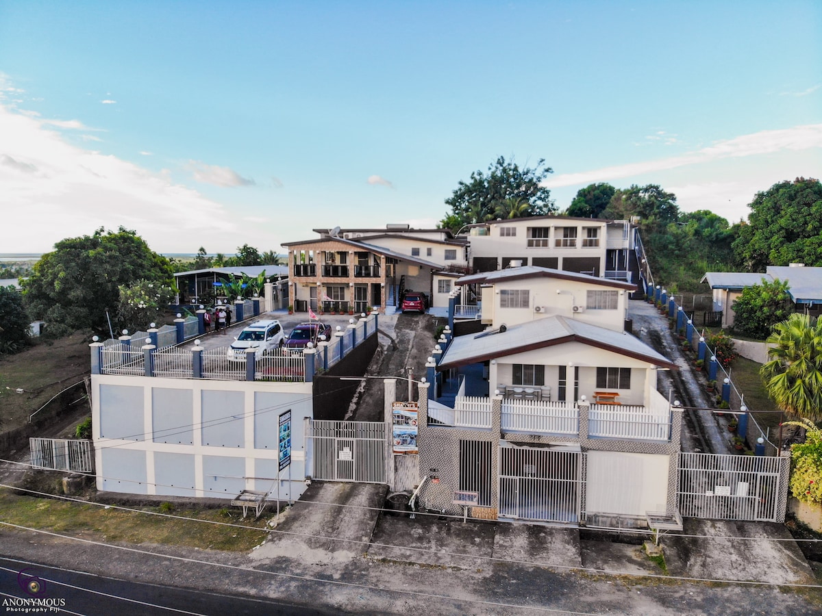 An aerial view showcases Shashi's Villa & Luxurious Apartments, highlighting the secured premises with ample parking space. The structure comprises multiple buildings surrounded by greenery and features electronically gated access. A clear blue sky adds to the overall ambiance of the well-kept environment.