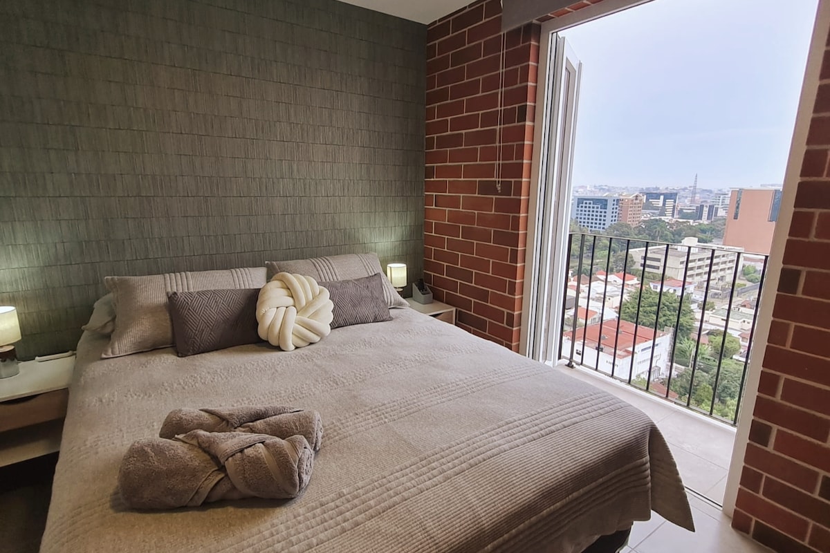 A spacious bed is positioned against a textured green wall, complemented by neutral-toned pillows. Natural light enters through an open balcony door, revealing a view of the city skyline. Soft linens and a towel sculpture add to the room's inviting appearance.