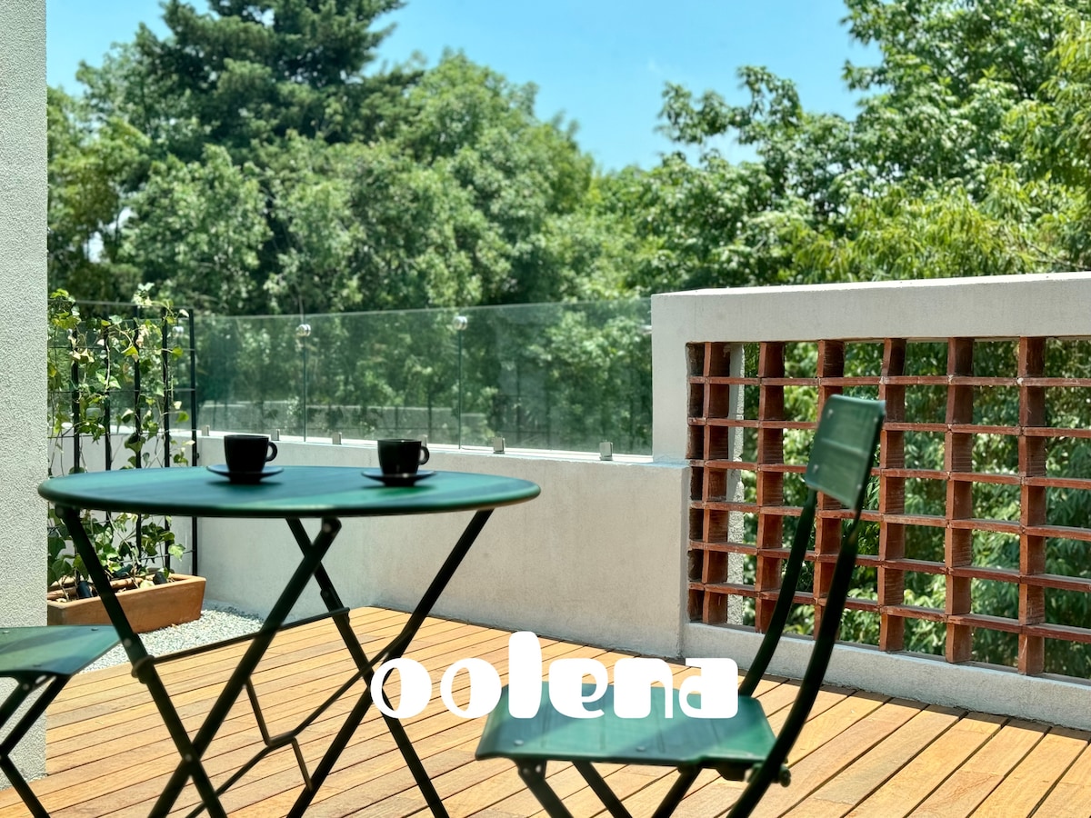 A private terrace features a small round table with two chairs, complemented by two coffee cups. Lush greenery surrounds the space, providing a serene atmosphere. Glass panels offer unobstructed views, enhancing the connection with the outdoors.