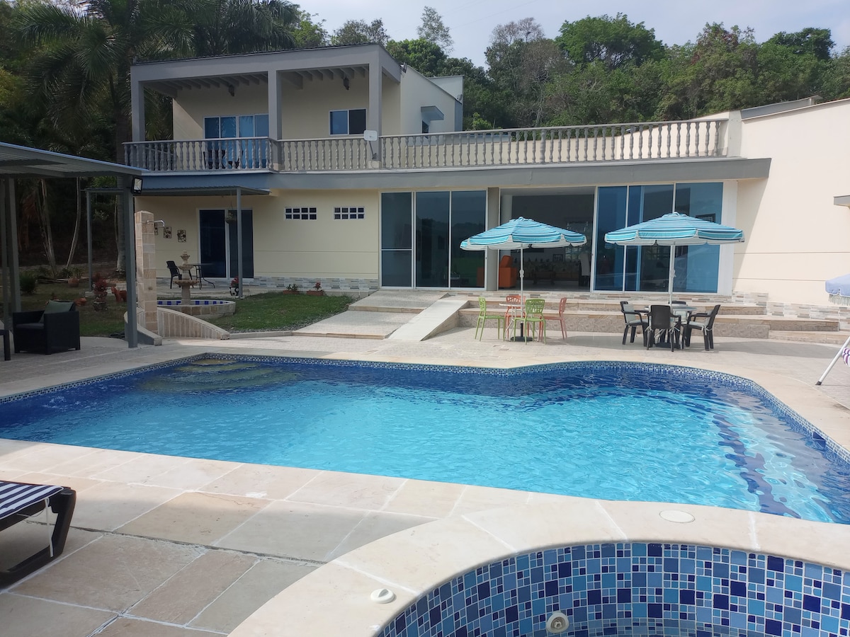 An inviting outdoor area features a clear blue pool surrounded by sun loungers and patio furniture, including tables with umbrellas. The spacious house is visible in the background, showcasing large windows that allow natural light to fill the interior.