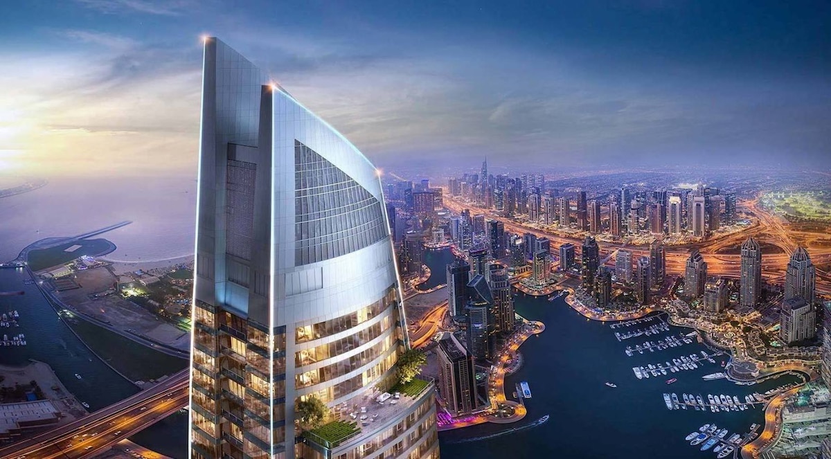 An aerial view captures the iconic tower of Dubai Marina, showcasing its modern architectural design. The vibrant city skyline and shimmering waterways extend into the distance, with a sunset casting soft light over the scene. The vast expanse of the sea is visible, enhancing the urban landscape.
