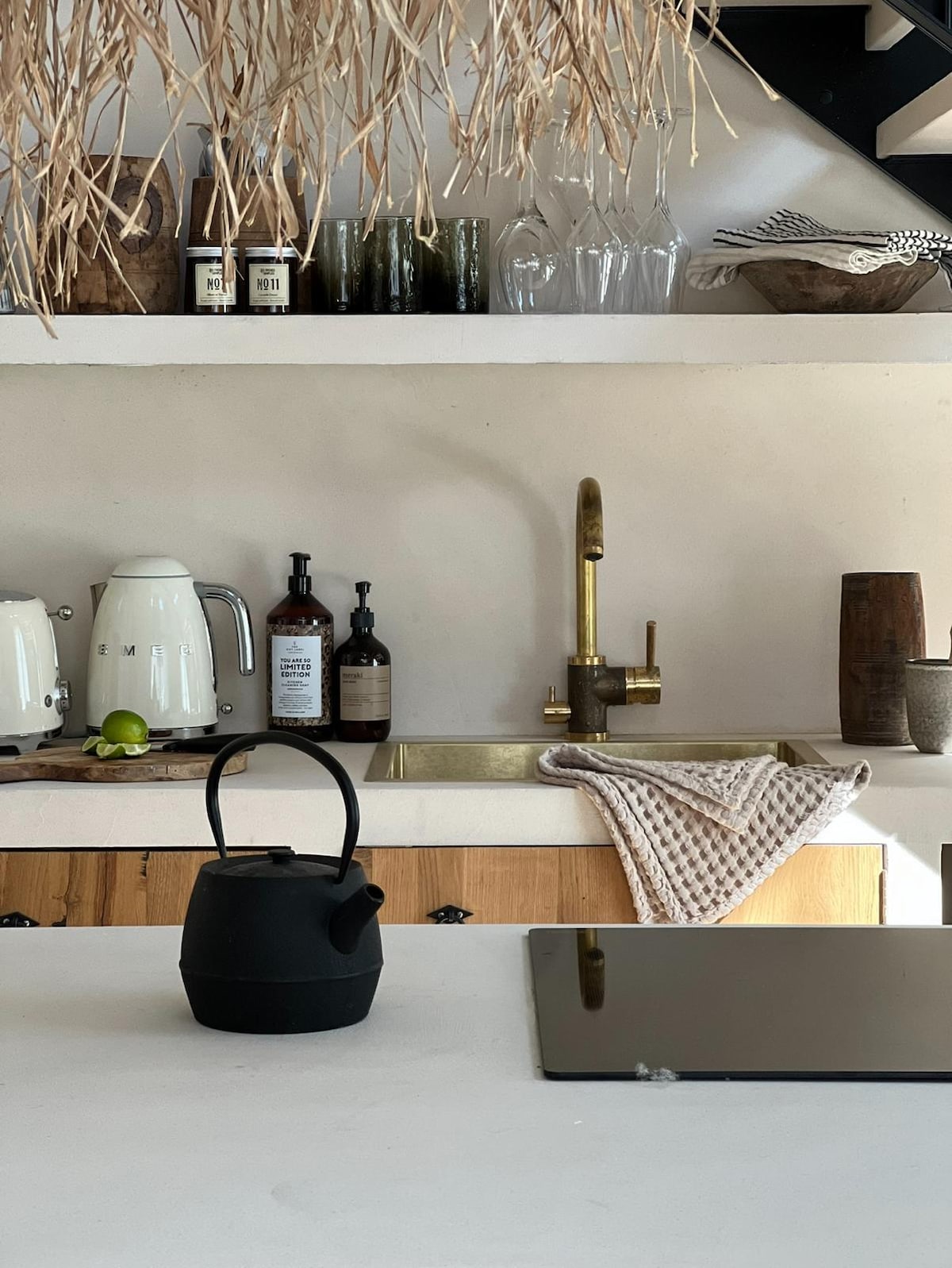 A minimalist kitchen setup highlights a black kettle resting on a smooth countertop. Behind it, a brass faucet is positioned above a sink, complemented by various kitchen items on open shelves, including glassware and bottles, creating a functional yet stylish cooking space.