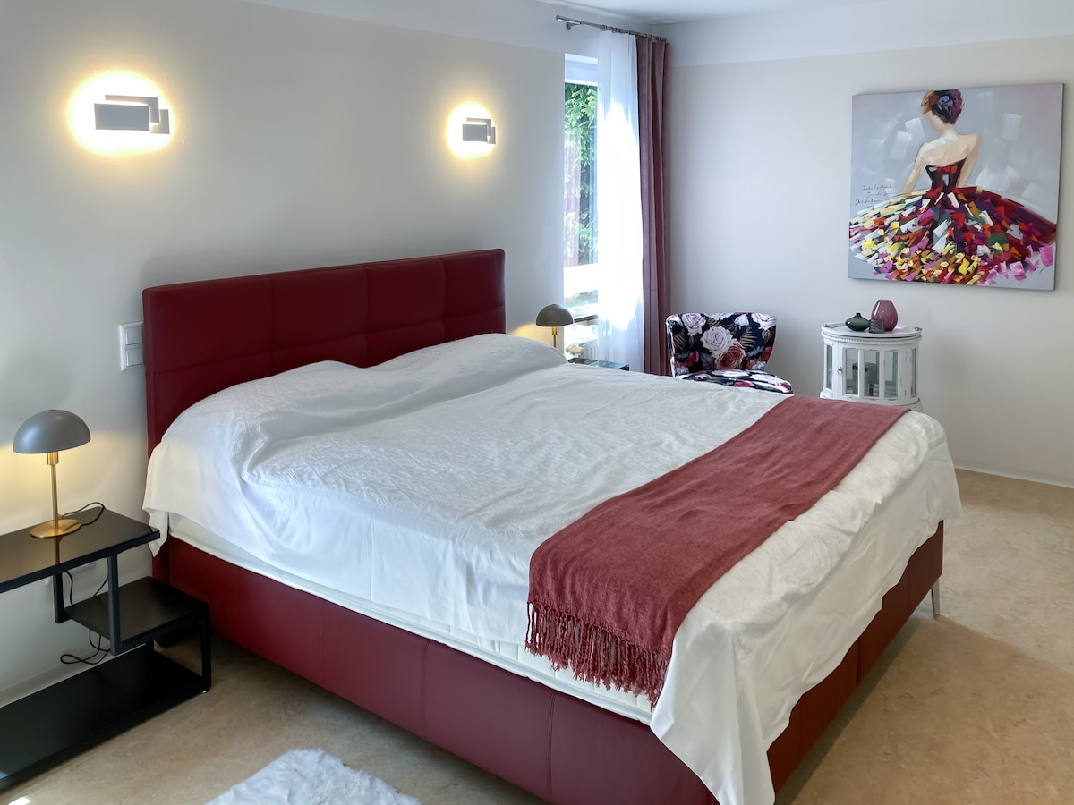 A spacious sleeping area is furnished with a large bed featuring an upholstered headboard. Soft lighting illuminates the room, complemented by a decorative painting and a cozy seating area. Natural light streams in through the window, enhancing the tranquil atmosphere.