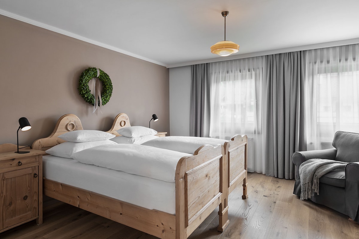 A serene bedroom features two wooden beds with white bedding and decorative pillows. A round green wreath hangs on the wall, complemented by soft gray curtains framing the windows. A comfortable gray armchair is situated in the corner, accompanied by a knitted throw blanket.