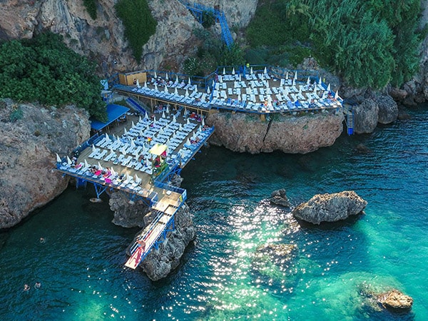 A seaside platform is depicted, featuring numerous lounge chairs arranged for relaxation. The platform extends over clear turquoise waters, with rocky formations visible nearby. Sunbathers can be seen enjoying the space, enhancing the inviting atmosphere of the beach setting.